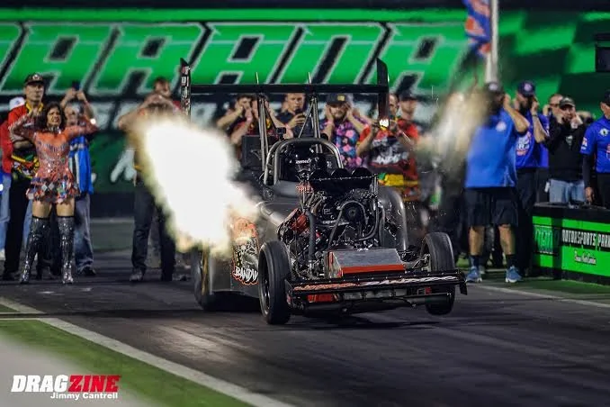 Ryan Hill's Nitro Bandido Fuel Altered at the starting line at night, with header flames and Mad Maxine beside the car